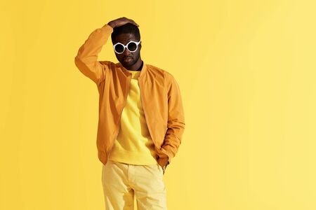 Fashion portrait of black man in stylish clothes and sunglasses on yellow background. Handsome african american male model in orange jacket posing in studioの写真素材