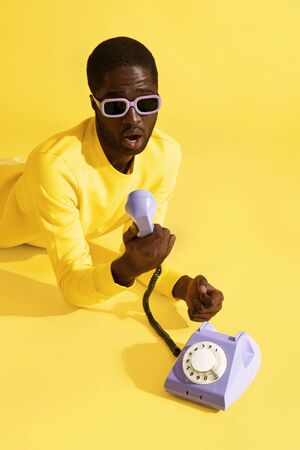 Surprised man in sunglasses with purple phone on yellow background. Portrait of shocked black male model with emotional scared face expression with telephone in studioの写真素材
