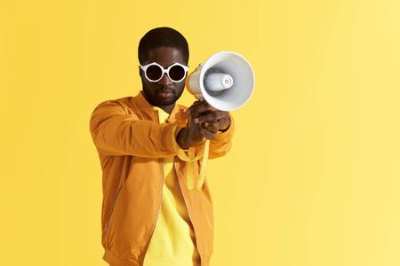 Black man in fashion sunglasses with megaphone portrait on yellow background. Male model in stylish clothes and eyewear with loud speaker in studioの写真素材