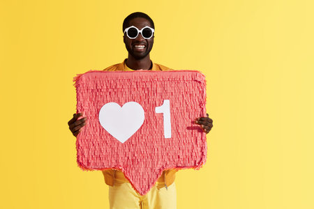 Like. Smiling black man with like icon, heart pinata on yellow background. Studio portrait of...
