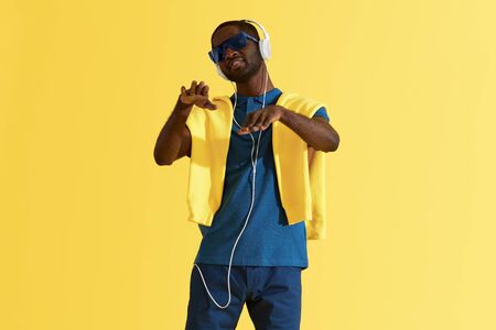 Man in headphones listening music and dancing on yellow background portrait. Happy black male model in headphone, sunglasses and fashion clothes in studioの写真素材