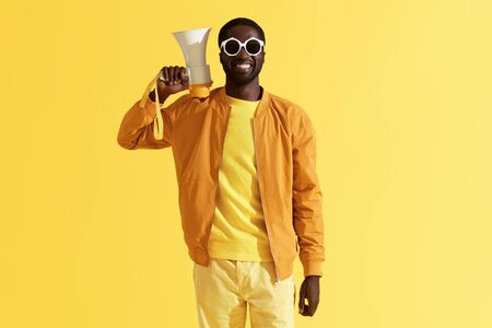 Fashion portrait of smiling black man with megaphone on yellow background. Happy young african american male model in stylish sunglasses with loud speaker in studioの写真素材