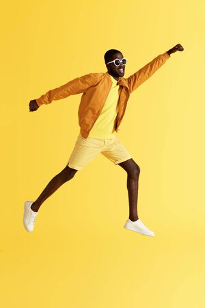 Jump. Man jumping in air flying like superhero on yellow background. Full length portrait of smiling black male model in fashion clothes and sunglasses having fun at studioの写真素材