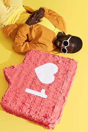 Black man lying with social media notification, like icon pinata on yellow background. Studio portrait of happy smiling male model relaxing near heart sign buttonの写真素材