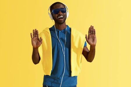 Man in headphones listening music and singing in studio portrait. Happy black male singer in headphone, stylish sunglasses and fashion clothes on yellow backgroundの写真素材