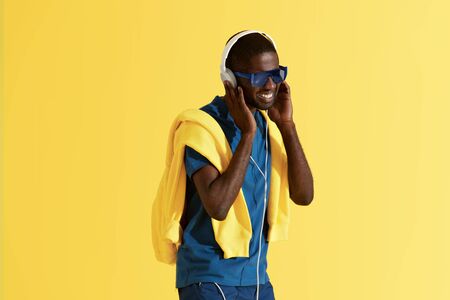 Fashion. Man listening music in headphones on yellow background. Colorful portrait of happy smiling black male model in headphone, fashion sunglasses and clothes at studioの写真素材