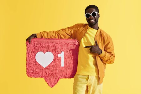 Like. Colorful portrait of happy black man with heart like icon pinata on yellow background. Studio shot of smiling male model pointing with finger on social media signの写真素材