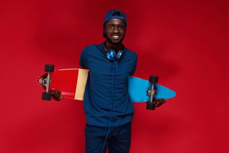 Fashion. Smiling african man in stylish outfit with skateboard at studio on red background. Happy black male model in blue cap, polo shirt with headphones on neck and skate boardの写真素材