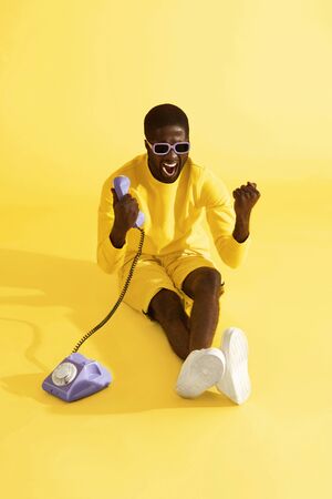 Happy excited man with phone screaming, celebrating on yellow background. Emotional black male model with retro purple telephone in studioの写真素材