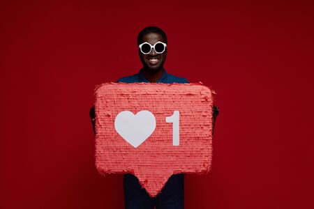 Happy black man with social media like icon pinata on red background. Portrait of smiling male model in sunglasses demonstrating huge heart notification signの写真素材