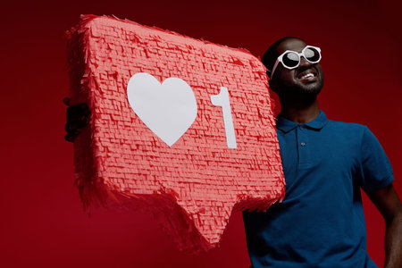 Happy black man with social media like icon pinata on red background. Portrait of smiling male model...