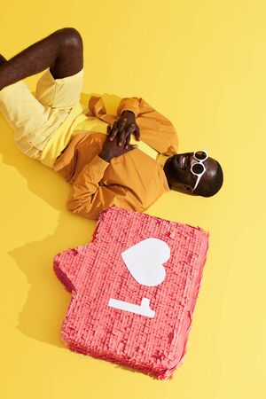 Black man lying with social media notification, like icon pinata on yellow background. Studio portrait of happy smiling male model relaxing near heart sign buttonの写真素材