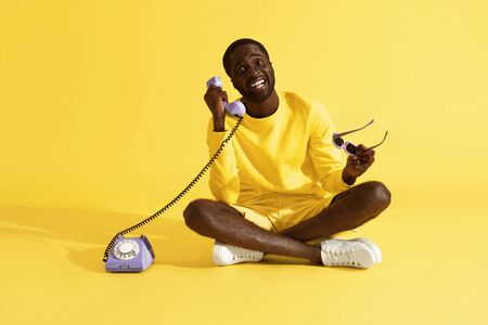 Happy man talking on purple phone sitting on yellow background. Smiling young african american male model in yellow clothes calling on violet retro corded telephone in studioの写真素材
