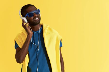 Fashion. Man listening music in headphones on yellow background. Colorful portrait of happy smiling black male model in headphone, fashion sunglasses and clothes at studioの写真素材