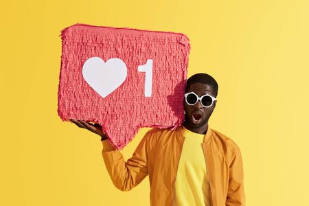 Social media like. Surprised black man with like icon pinata on yellow background. Colorful portrait of shocked male model in sunglasses with huge like signの写真素材