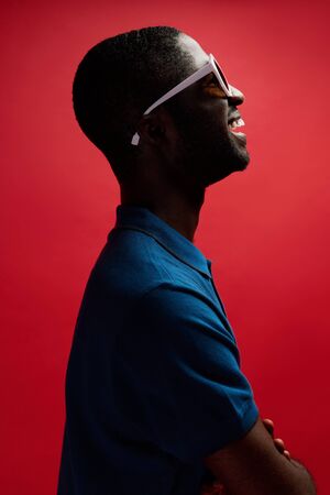 Fashion. Smiling black man in sunglasses on red background. Profile portrait of happy cheerful african american male model posing in shadows at studioの写真素材