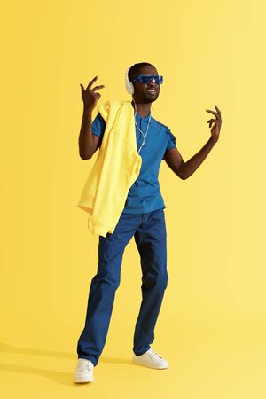 Man in headphones listening music, dancing in studio on yellow background. Full length portrait of happy black male model in sunglasses and fashion clothes enjoying soundの写真素材