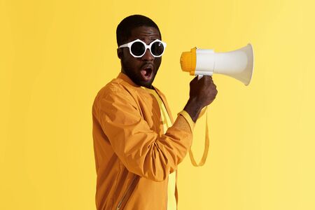 Surprised black man with megaphone on yellow background. Studio portrait of shocked african american male model in fashion sunglasses with loud speakerの写真素材