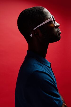 Fashion. Black man in sunglasses on dark red background. Profile portrait of handsome african american male model posing in shadows at studioの写真素材
