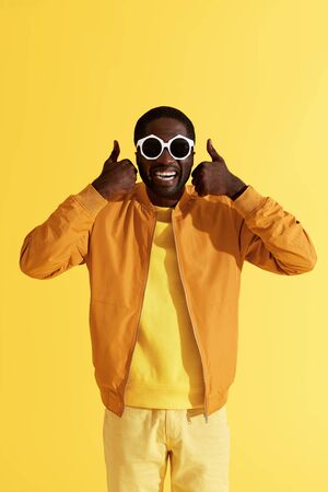 Smiling man in fashion sunglasses showing thumbs up on yellow background. Studio portrait of happy african american male model in colorful trendy clothes showing like with handsの写真素材