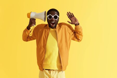 Happy black man with megaphone having fun, studio portrait. Surprised african american male model in fashion clothes and sunglasses with hands up on yellow backgroundの写真素材
