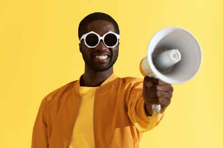 Smiling black man in fashion sunglasses with megaphone portrait on yellow background. Happy young...