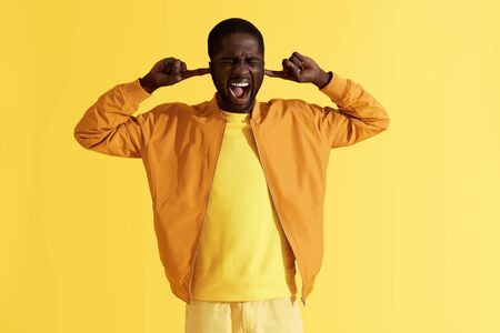 Stress. Man closing ears screaming loud on yellow background. Portrait of black male model sticking fingers in ears, shouting and ignoring noise, studio shotの写真素材