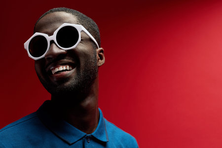 Fashion. Smiling black man in sunglasses on red background. Contrast portrait of happy cheerful...