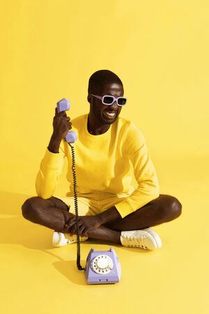 Man having stressful phone call sitting on yellow background. Male model with unhappy face having unpleasant talk on purple telephone in studioの写真素材