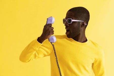 Surprised man in sunglasses with purple phone on yellow background. Portrait of shocked black male model with emotional scared face expression with telephone in studioの写真素材