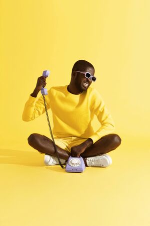 Man having stressful phone call sitting on yellow background. Male model with unhappy face having unpleasant talk on purple telephone in studioの写真素材