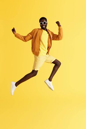 Jump. Happy man jumping in air on yellow background. Full length portrait of cheerful smiling black male model in fashion clothes and sunglasses having fun, studio shotの写真素材