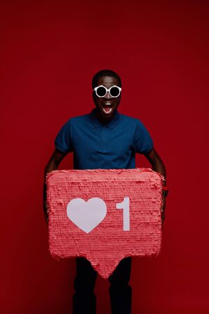 Happy black man with social media like icon pinata on red background. Portrait of smiling male model in sunglasses demonstrating huge heart notification signの写真素材