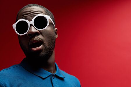 Surprised black man in fashion sunglasses on red background. Contrast portrait of shocked african american male model in stylish glasses and " wow " emotion at studioの写真素材