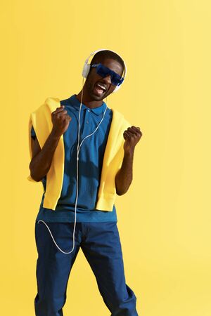 Man in headphones listening music and singing in studio portrait. Happy black male singer in headphone, stylish sunglasses and fashion clothes on yellow backgroundの写真素材