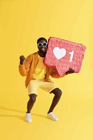 Smiling man with like icon, heart pinata on yellow background. Happy male with huge "like" sign button, social media symbol enjoying followers feedbackの写真素材