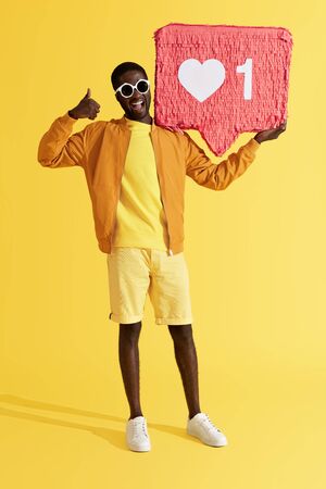 Smiling black man with like icon pinata showing thumbs up on yellow background. Studio shot of happy male model showing like with hand, holding heart button, social media symbolの写真素材