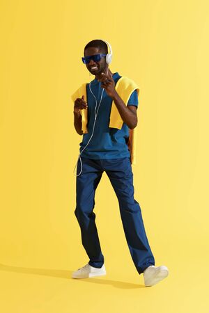 Man in headphones listening music, dancing in studio on yellow background. Full length portrait of happy black male model in sunglasses and fashion clothes enjoying soundの写真素材