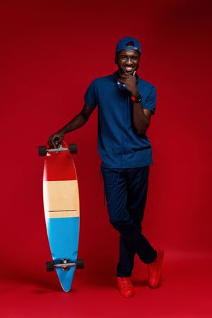 Fashion. Smiling african man in stylish outfit with skateboard at studio on red background. Happy black male model in blue cap, polo shirt with headphones on neck and skate boardの写真素材