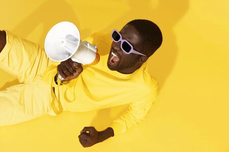 Man screaming in megaphone on yellow background. Portrait of african american male model in fashion wear using loud speaker in studioの写真素材