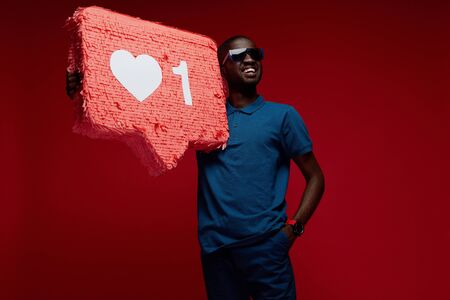 Happy black man with social media like icon pinata on red background. Portrait of smiling male model in sunglasses demonstrating huge heart notification signの写真素材