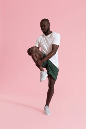 Man in sportswear stretching legs, warming up on pink background. Full length portrait of black male model in sport clothes doing leg stretches at studioの写真素材