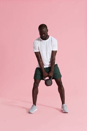 Workout. Man doing kettlebell swing exercise, sports training on pink background. Fitness male model exercising with kettle bell at studioの写真素材