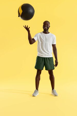 Sports man in sports wear with med ball on yellow background. Full length portrait of happy smiling male trainer in active wear with weight ball at studioの写真素材