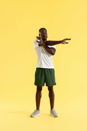 Workout. Man stretching arm, warming up on yellow background. Full length portrait of sporty male model in stylish sport wear doing arms stretch exercise at studioの写真素材