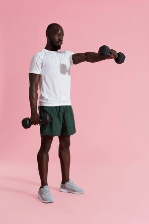 Man exercising, doing dumbbell front raise exercise on pink background. Fitness male model doing delta shoulders workout in gymの写真素材