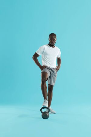 Sports man in sports wear with kettlebell on blue background. Full length portrait of african american male trainer in stylish active wear with weights at studioの写真素材