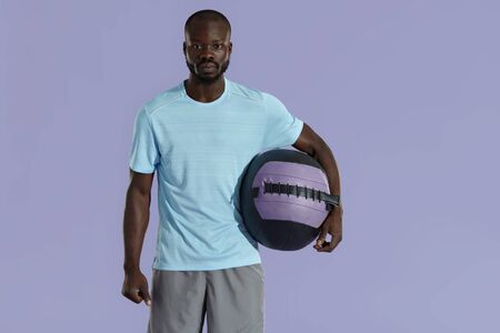 Sport man in fashion sportswear with med ball studio portrait. African american male model in fashionable active clothing with ball on purple color backgroundの写真素材