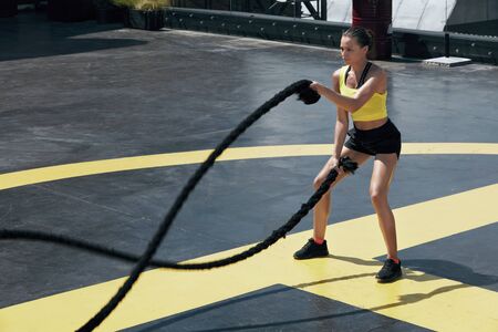 Sport woman doing battle ropes exercise workout at gym. Athletic girl exercising, doing functional fitness training with heavy ropes outdoors at streetの写真素材