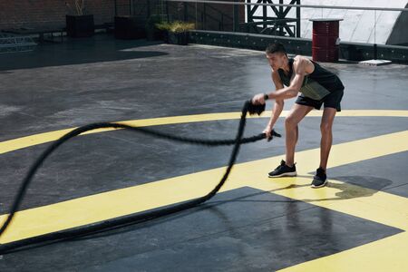 Sport man doing battle ropes exercise workout at gym. Athletic male exercising, doing functional fitness training with heavy ropes outdoors at streetの写真素材